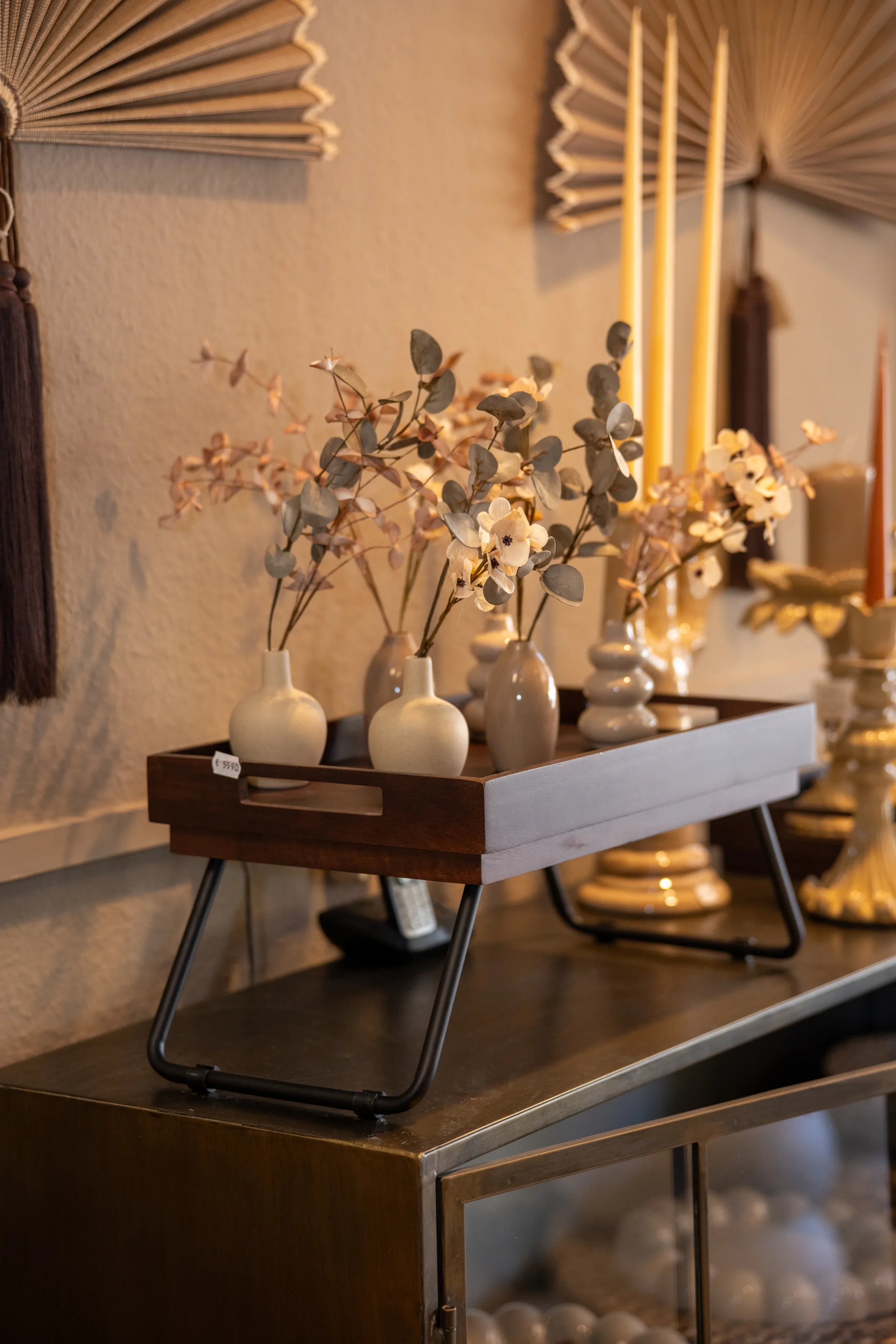 Holztablett mit kleinen Vasen und Zweigen auf einem Sideboard, dahinter Spitzkerzen und Wandfächer