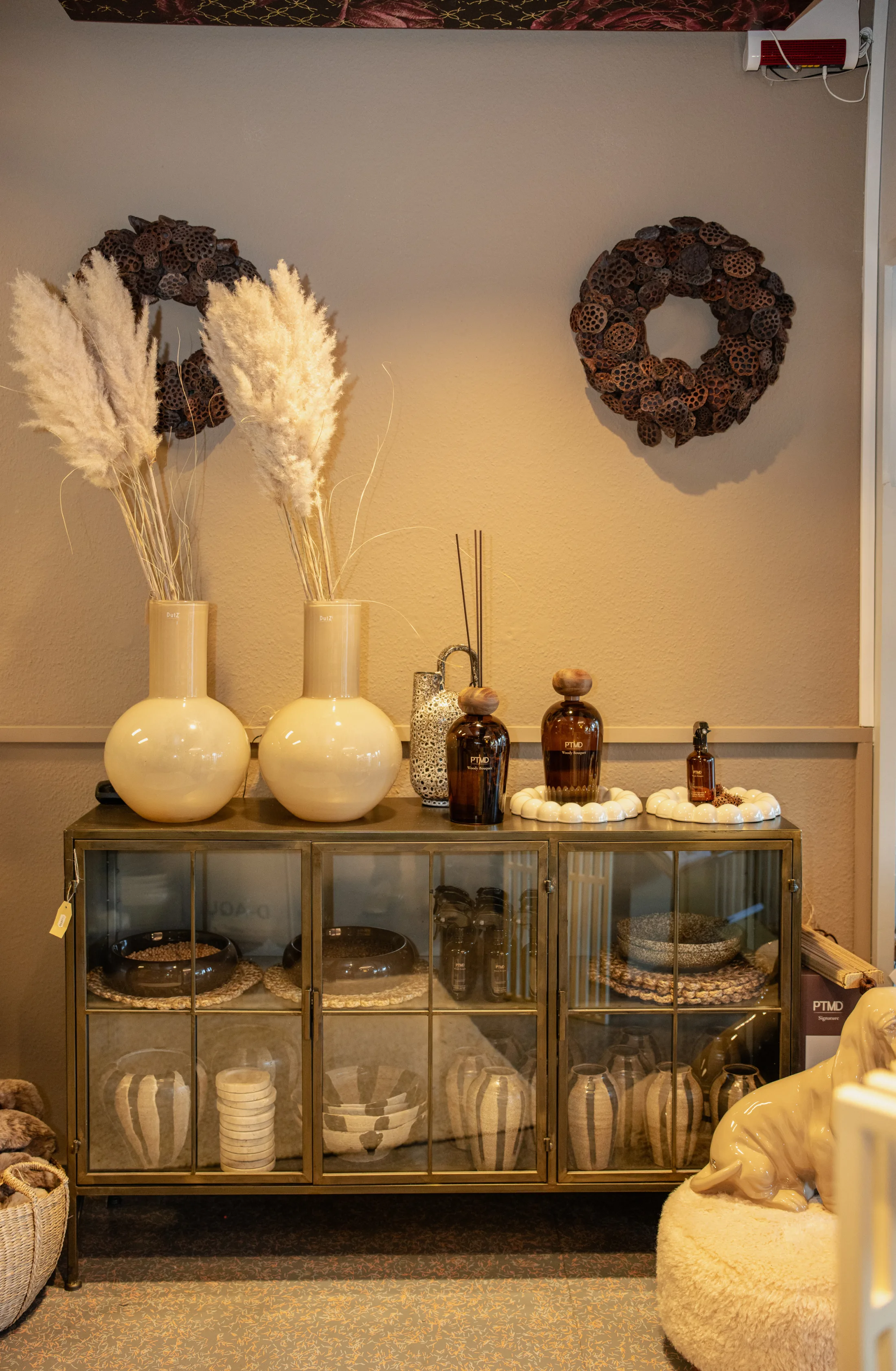 Sideboard mit Glastüren, darauf zwei bauchige Vasen mit Pampasgras, Duftflakons und Kränze aus Lotuskapseln an der Wand